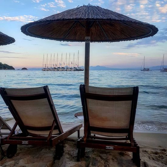 Due sedie a sdraio sotto un ombrellone di paglia, di fronte al mare con barche a vela e una grande nave al tramonto.