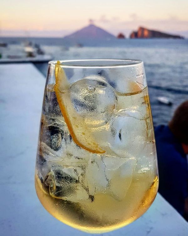 Cocktail con guarnizione di limone e ghiaccio, sfondo oceano, tramonto sfocato.