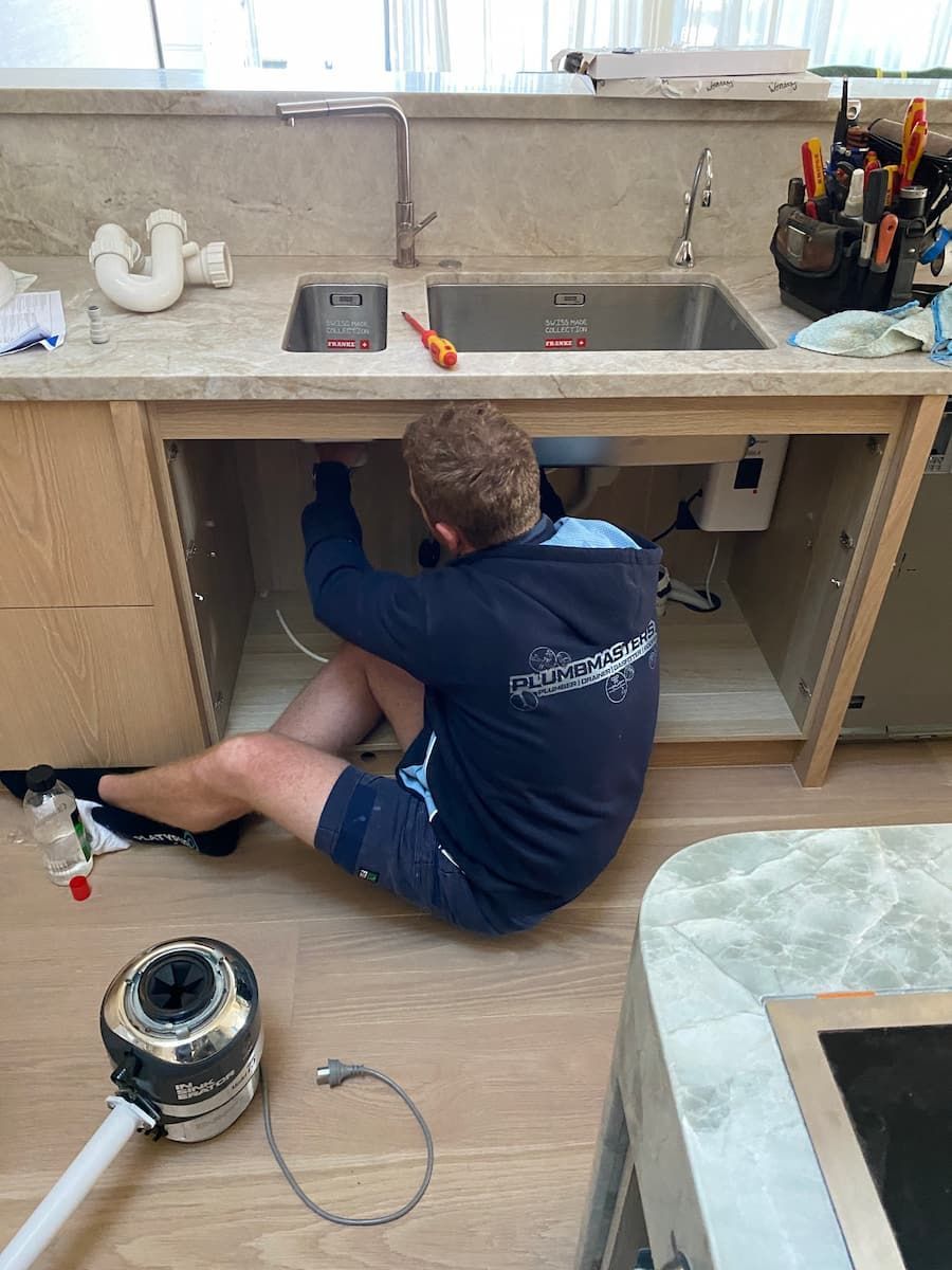 A Man Is Kneeling Under A Sink In A Kitchen — Plumbmasters In Nerang, QLD