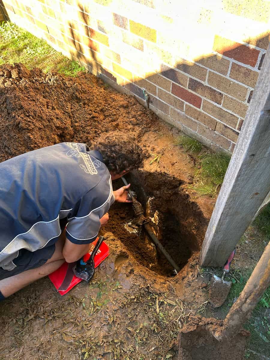 A Man Is Digging A Hole In The Ground Next To A Brick Wall — Plumbmasters In Tweed Heads, NSW