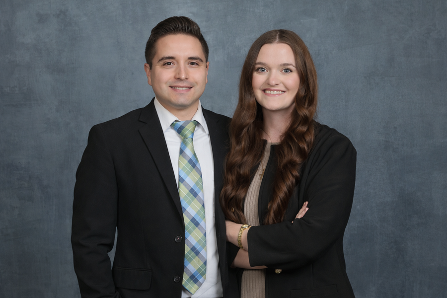 Two people in business attire standing close together, smiling. Gray background.