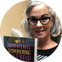 A woman wearing glasses is holding a book titled shining shimmering blended