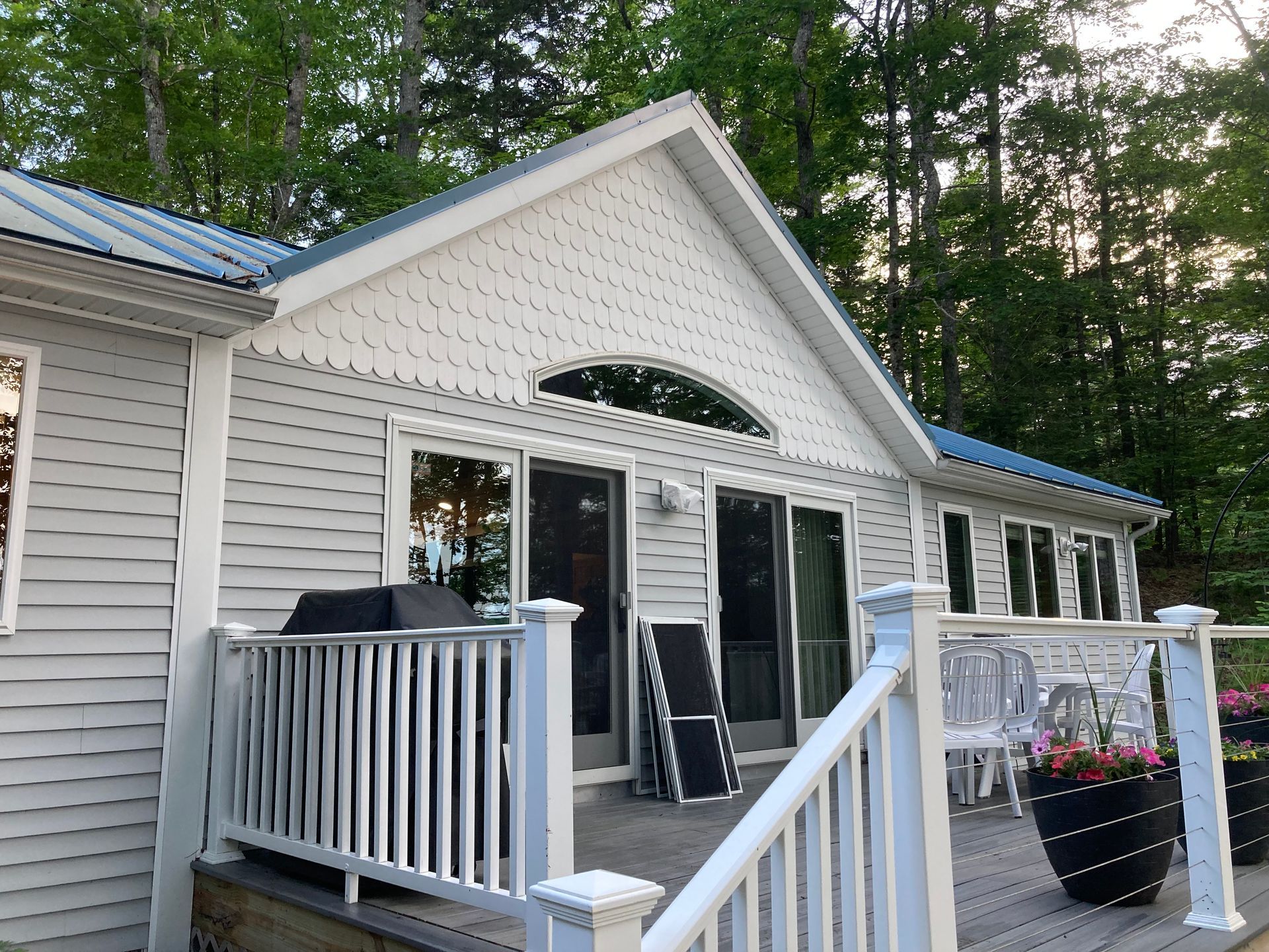 White house deck with sliding glass doors, a grill, and potted flowers beside a wooded backyard