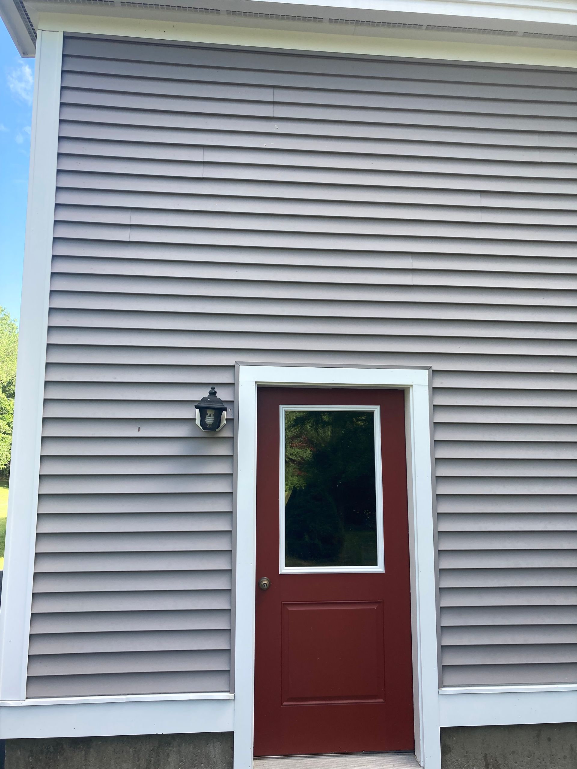 Red front door on gray siding with a small window and black wall lantern
