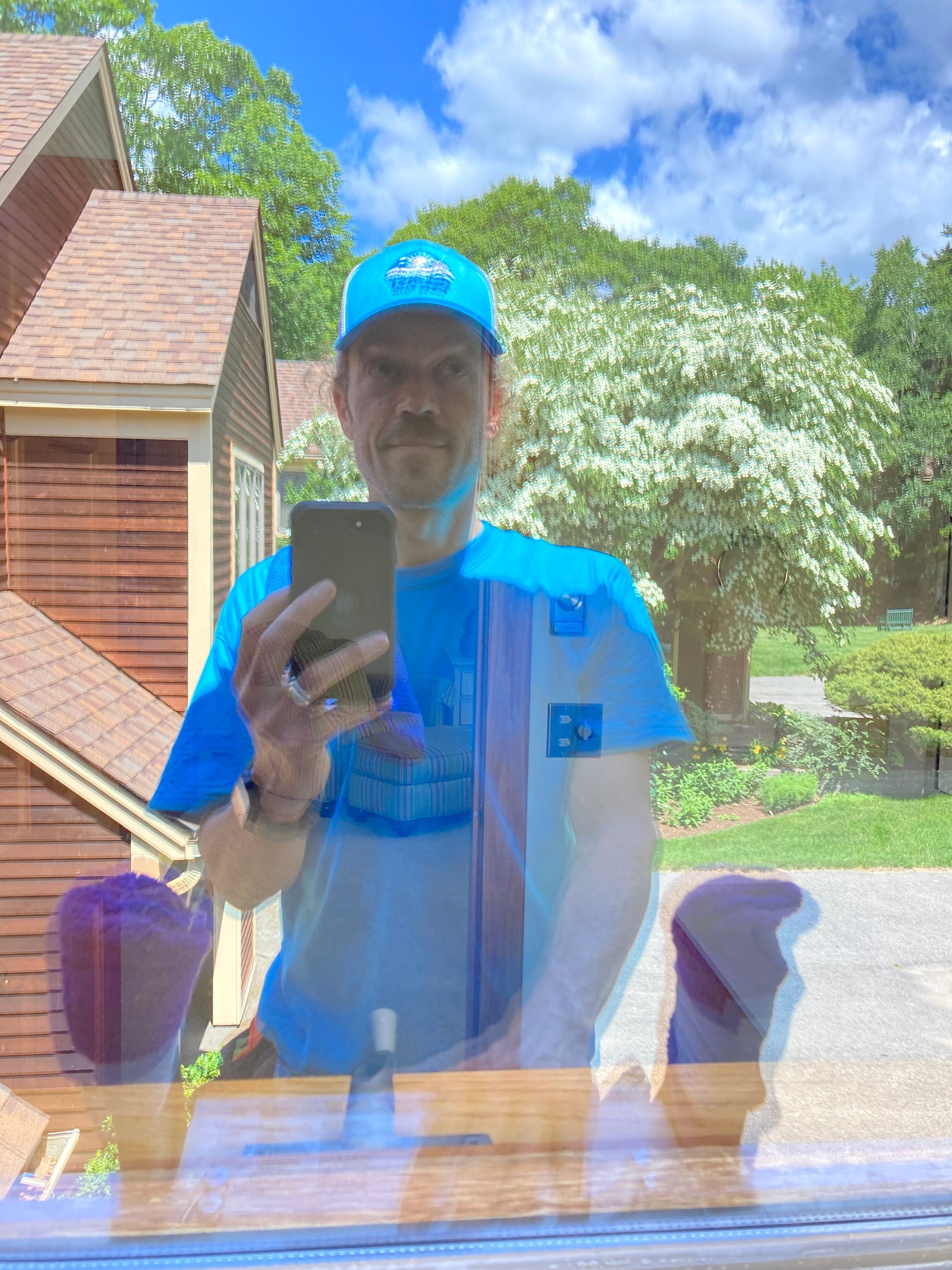 Person in blue shirt and cap taking a mirror selfie outdoors by a house and garden.