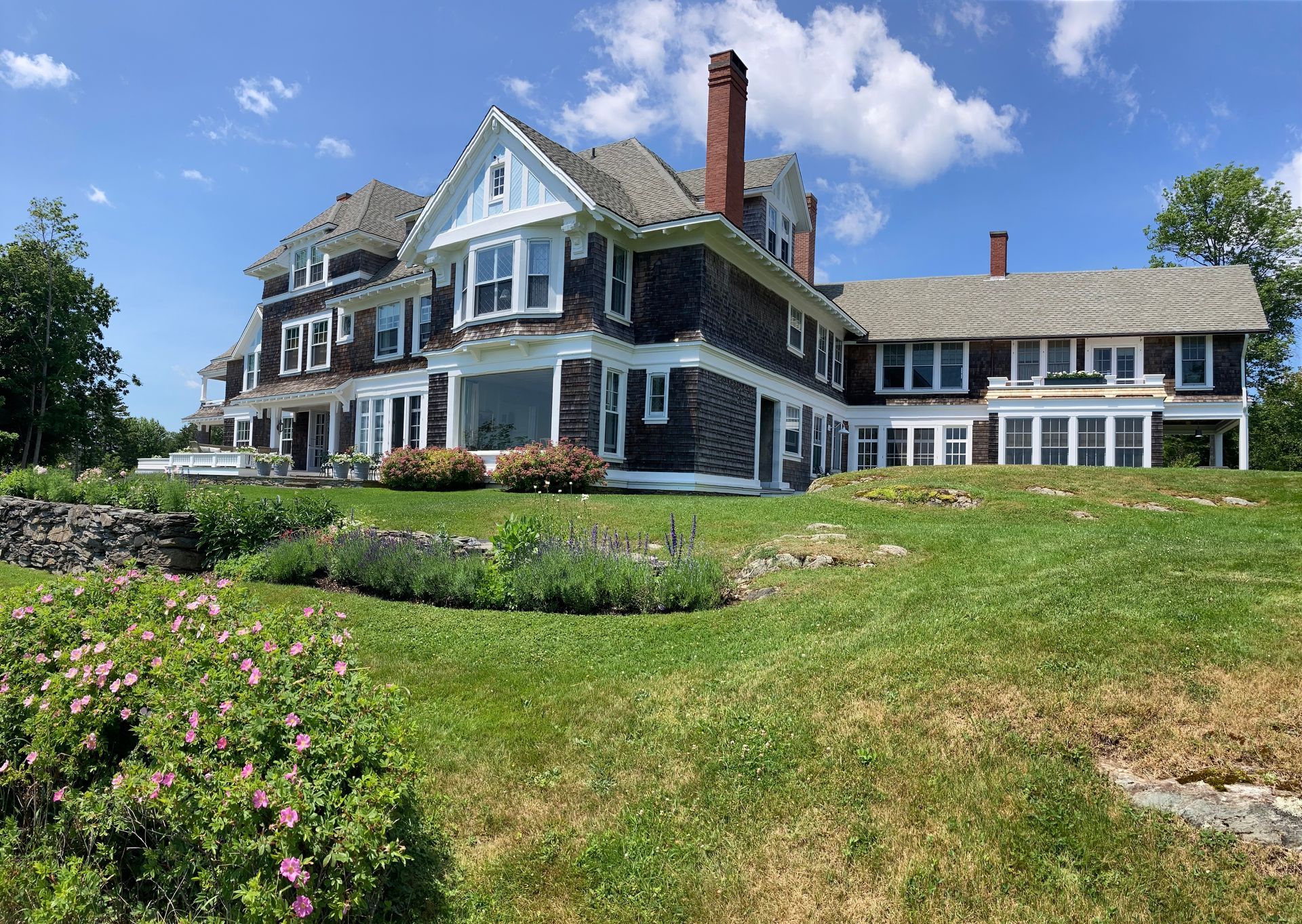 Large gray seaside house with white trim, stone wall, and manicured lawn under a blue sky