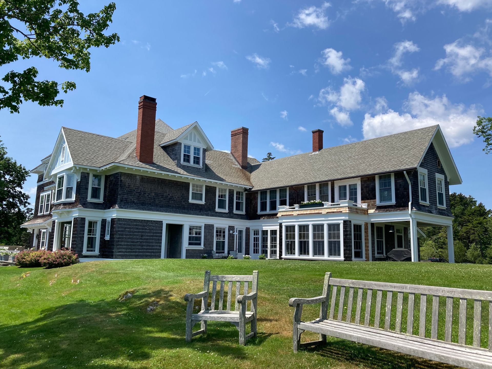 Large dark-blue seaside house with white trim, on a green lawn under a blue sky, with benches in front