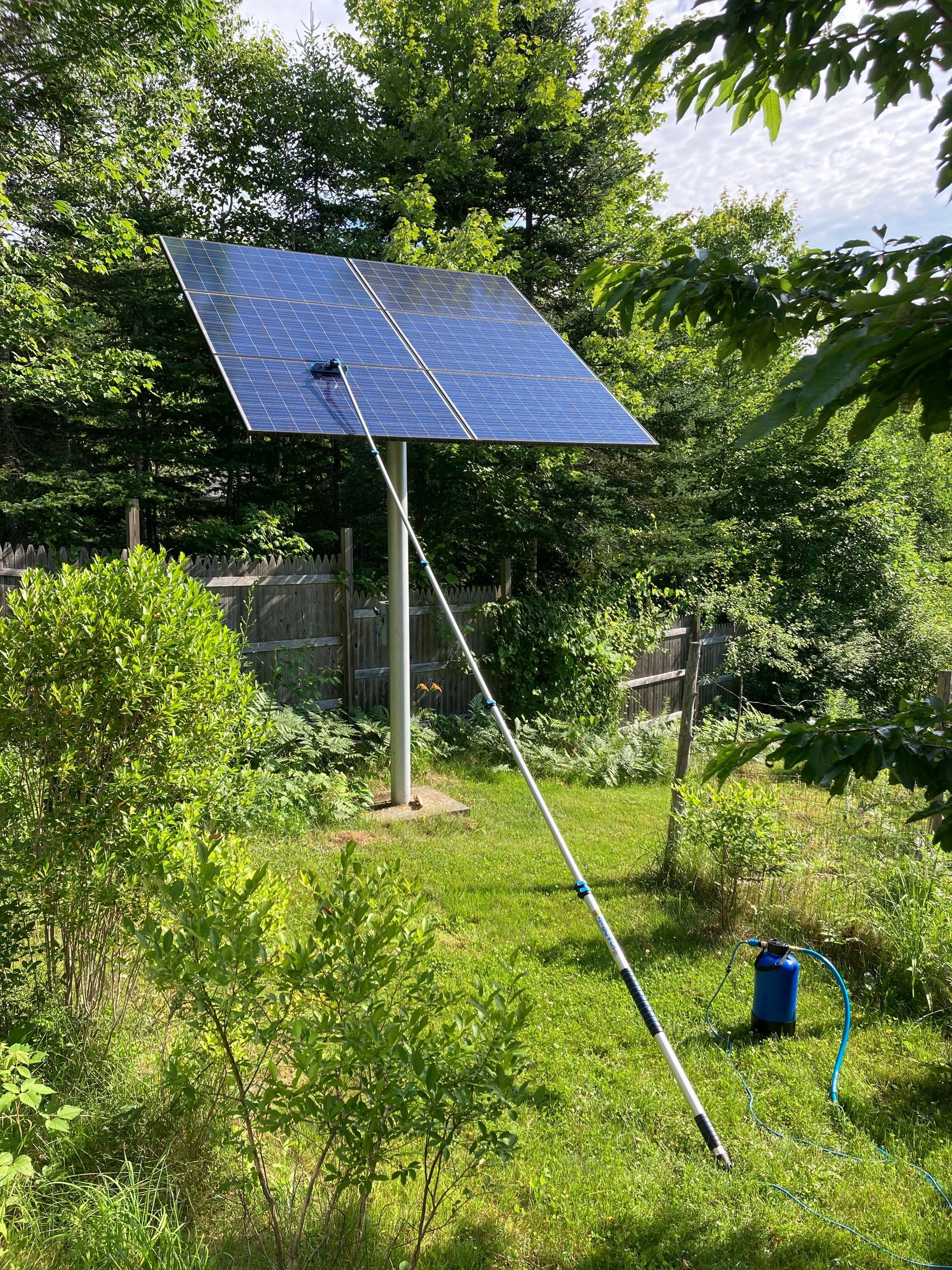 Blue solar panel on a metal pole in a green garden, with a support rod and hose nearby.