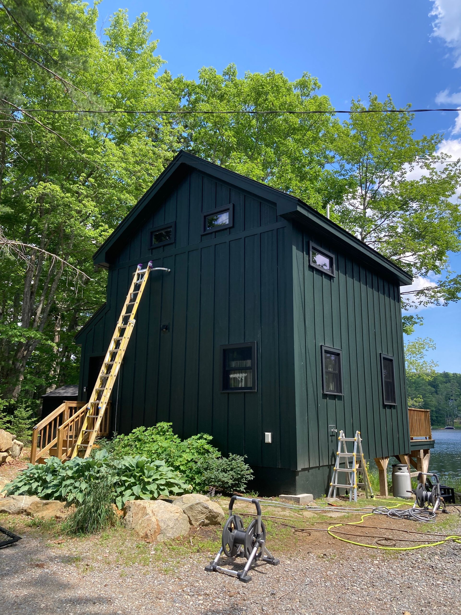 Small dark green cabin with a ladder and chair in a leafy yard under a bright blue sky