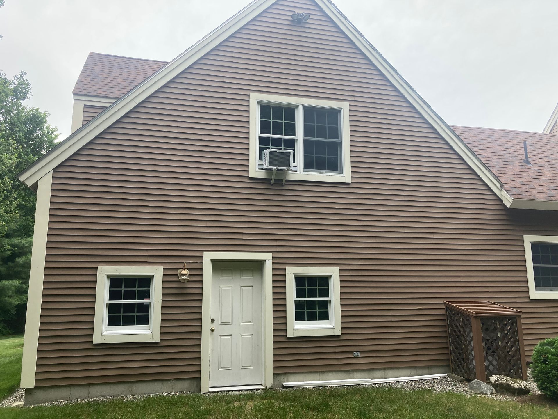 Brown-sided house exterior with white trim, a centered door, and a second-story window air conditioner.