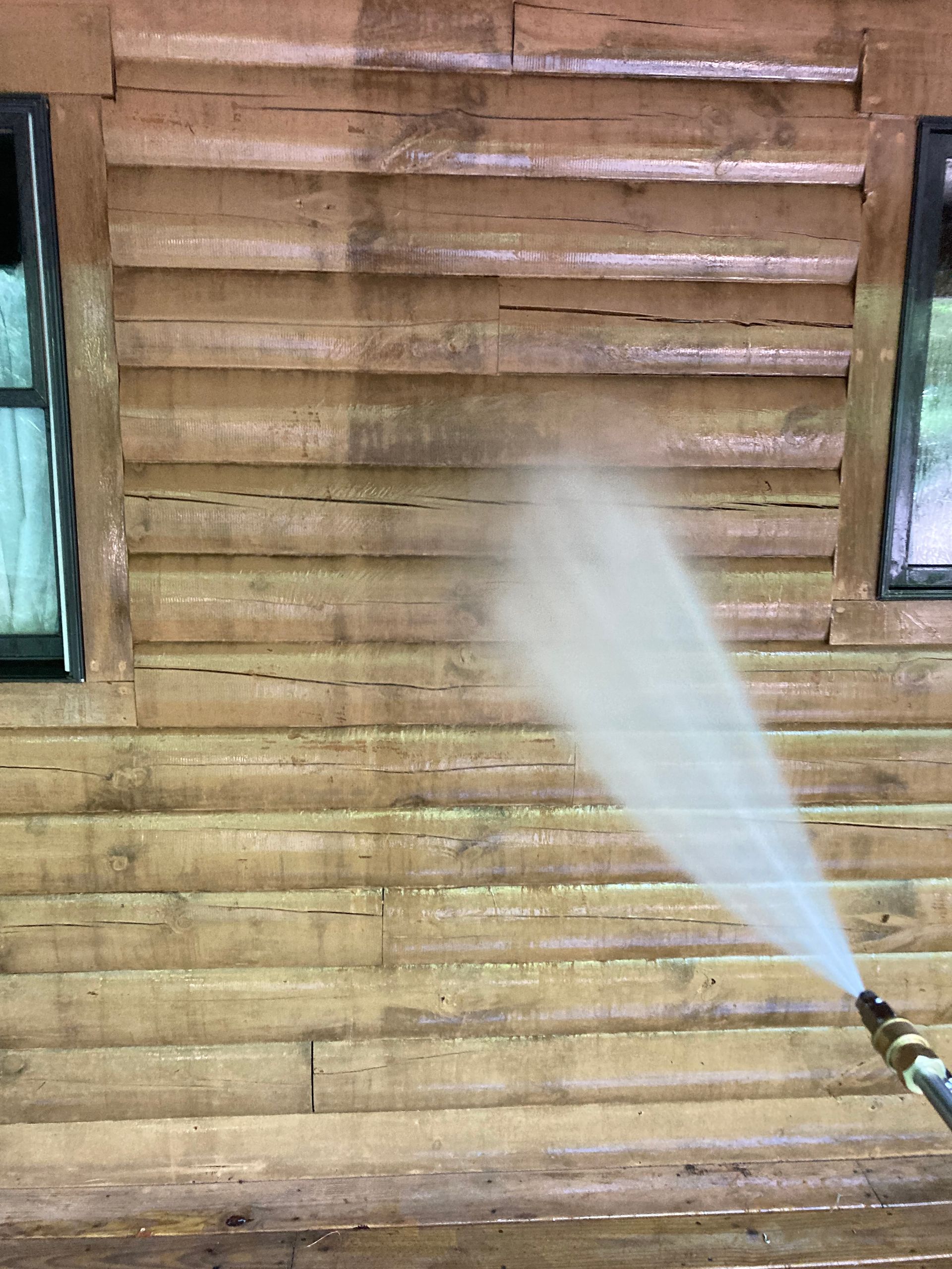 Person pressure-washing a wooden wall, spraying water on stained horizontal boards.