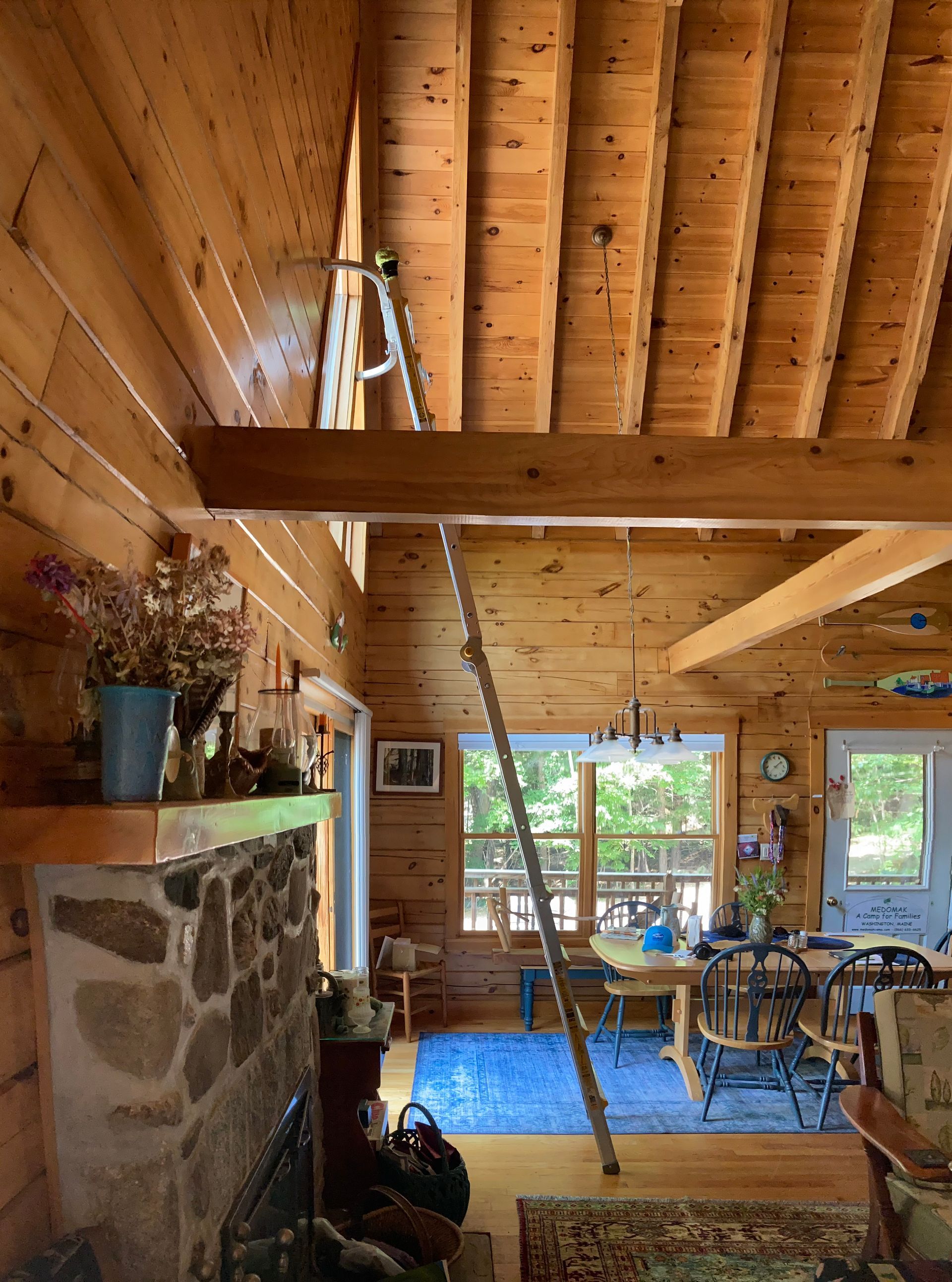 Cozy log cabin interior with wood beam ceiling, stone fireplace, loft ladder, and dining area by the windows