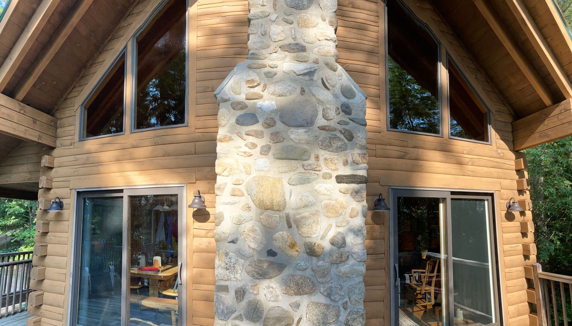 Rustic log cabin facade with a central stone chimney, tall windows, and a peaked wooden roof.