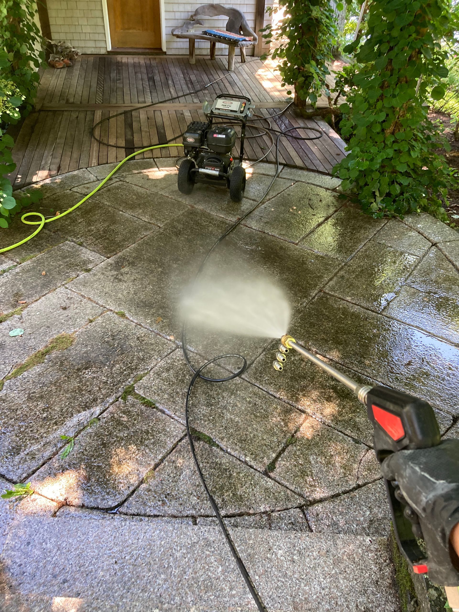 Person pressure-washing a wet stone patio beside a house and hedges