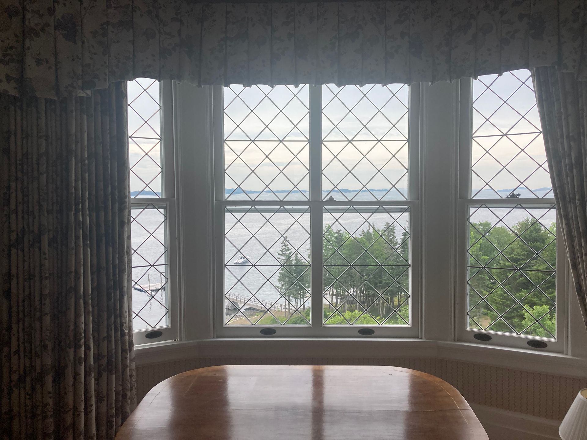 Bay window with diamond-pane glass overlooking the sea, framed by curtains and a wooden table.