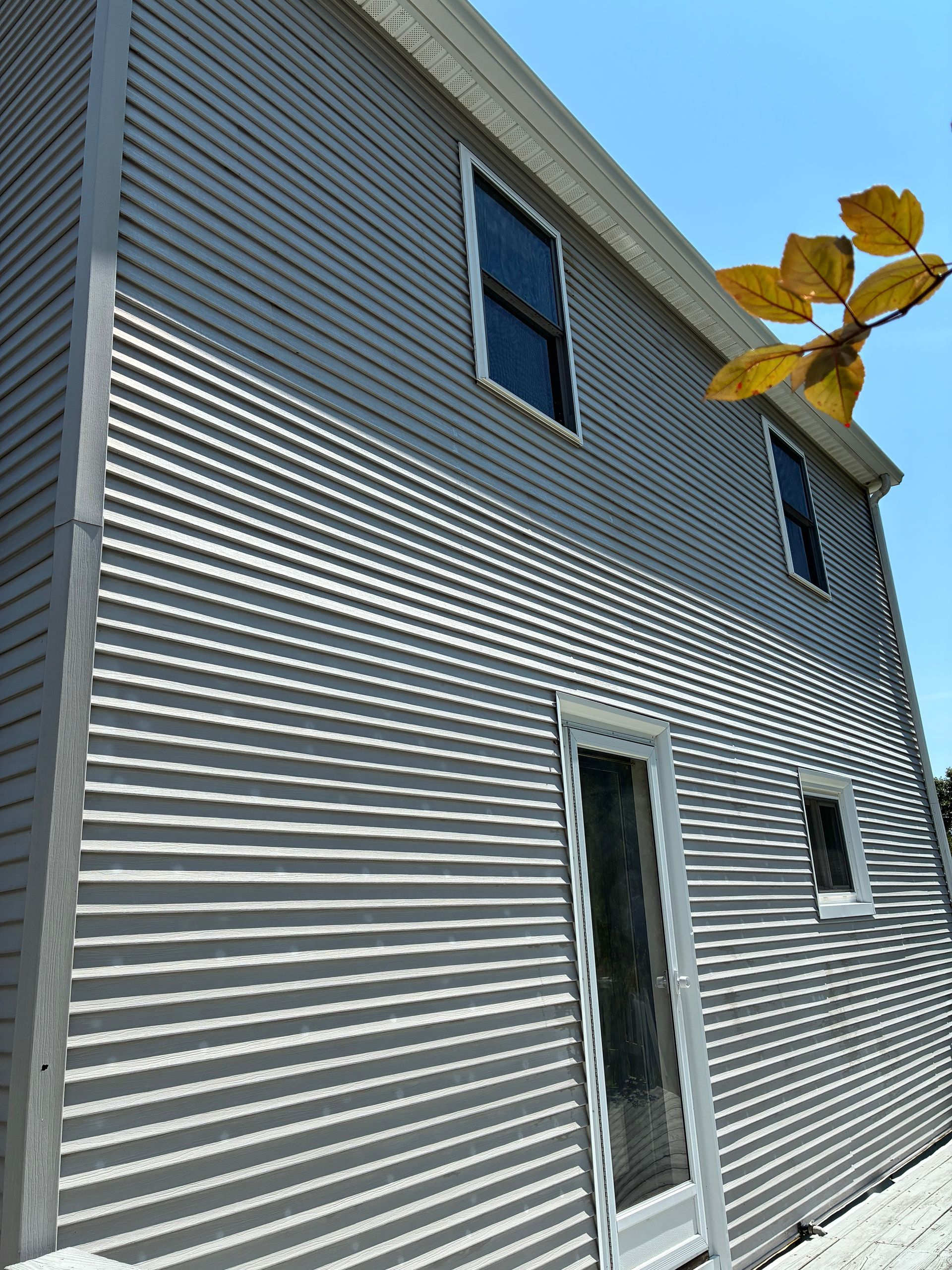 Gray siding on a two-story house with white trim and a glass door, viewed from below against blue sky.