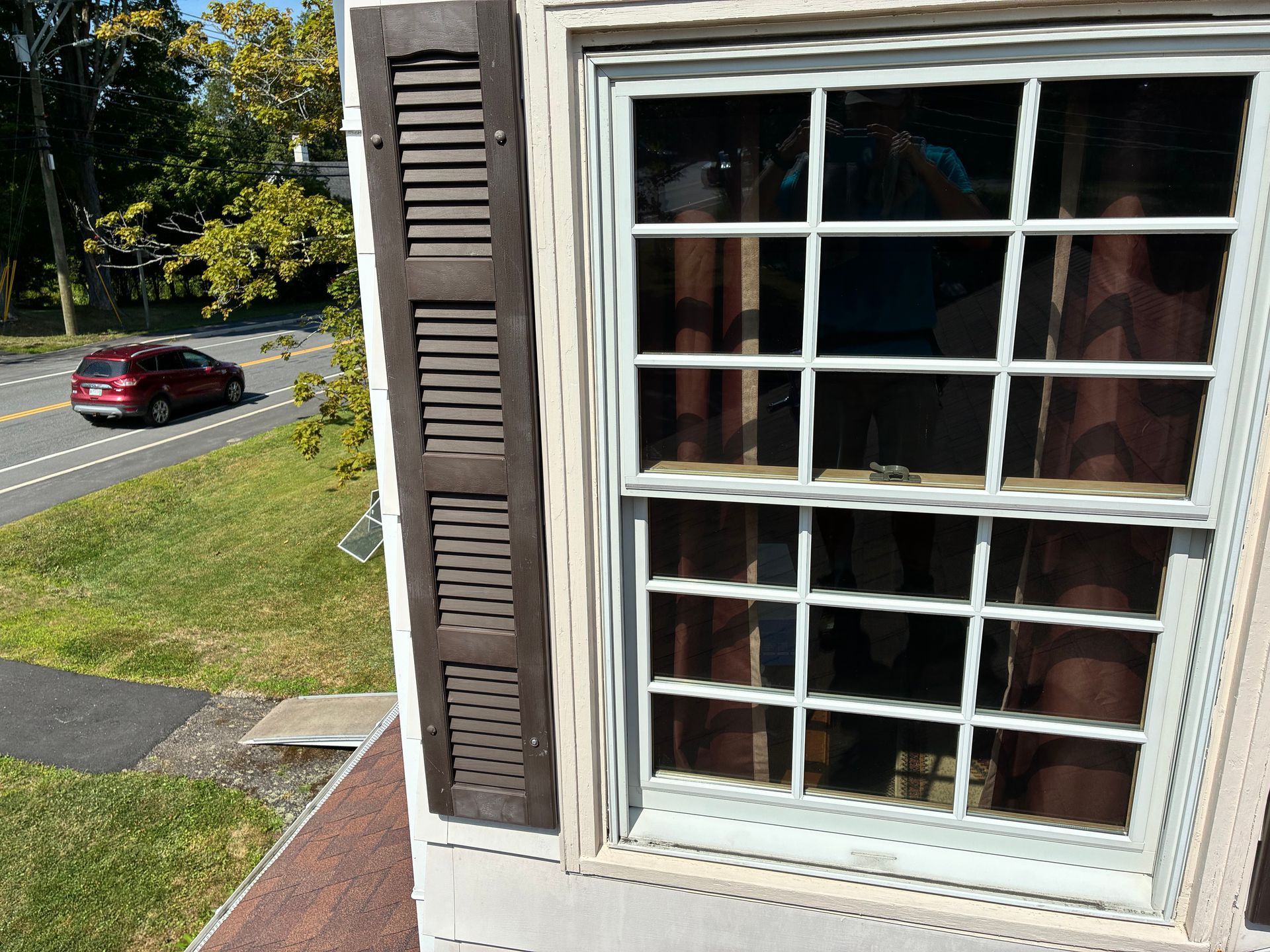 House window with white trim and brown shutters, overlooking a street with a red car and grassy yard