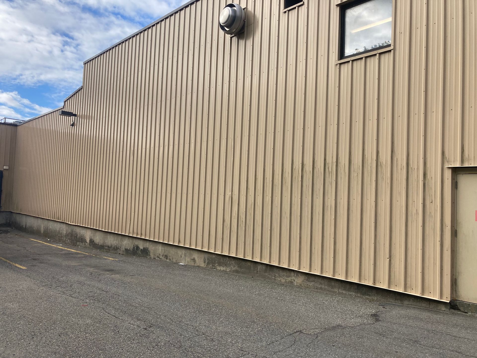 Tan corrugated metal building wall beside an empty paved lot under a partly cloudy sky
