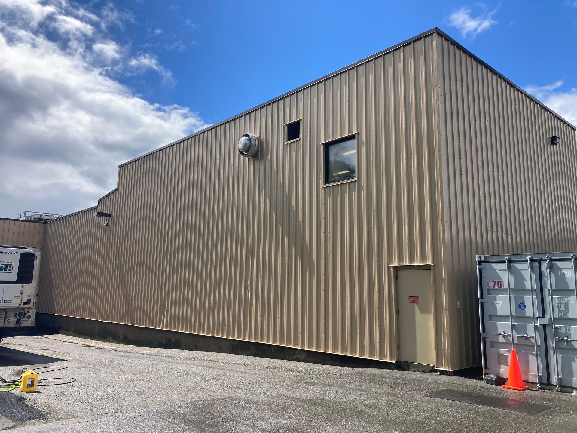 Tan metal warehouse building with gravel lot, small door, window, and orange traffic cone under a blue sky