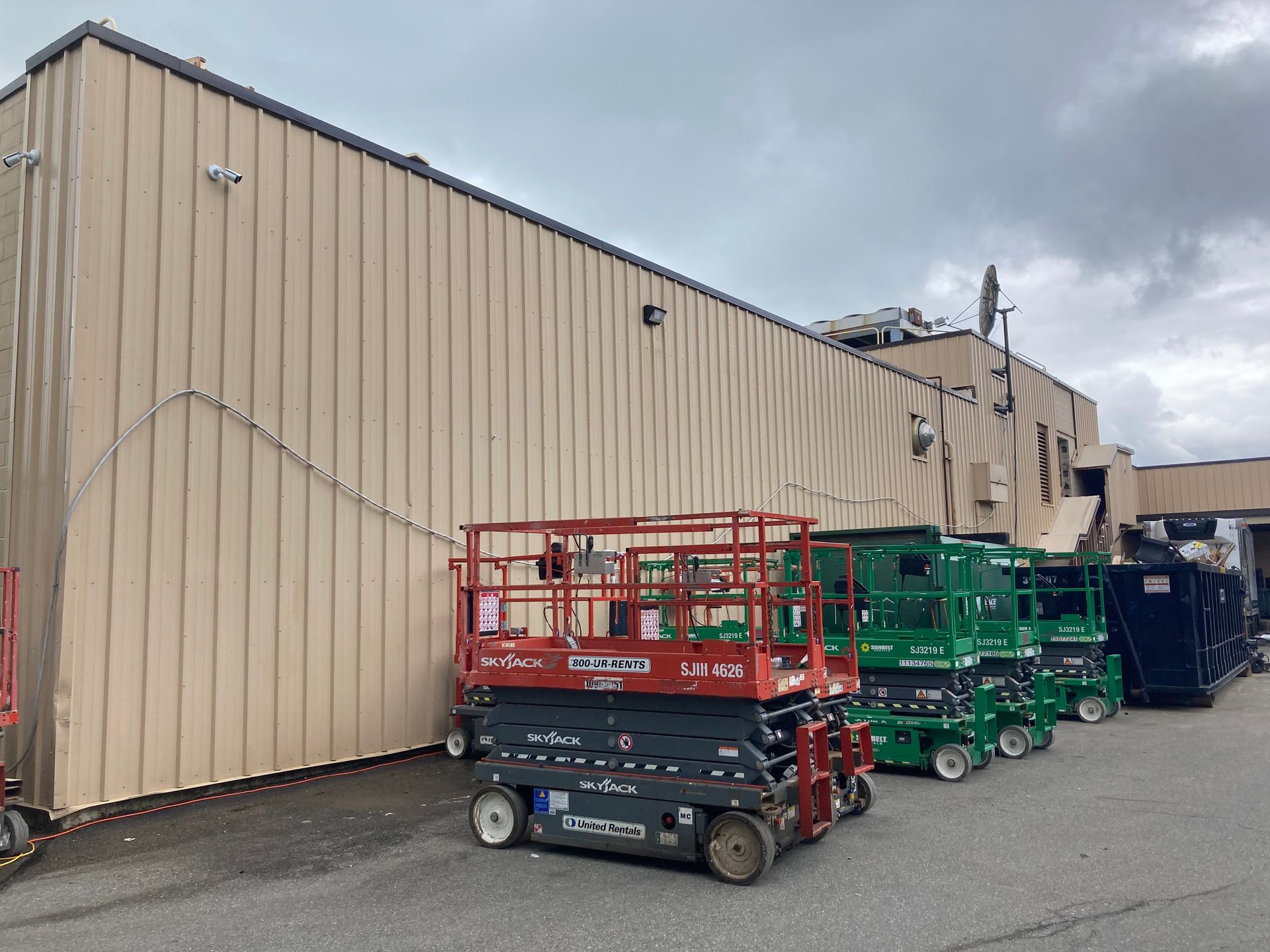 Line of red and green scissor lifts parked beside a beige industrial building under cloudy skies