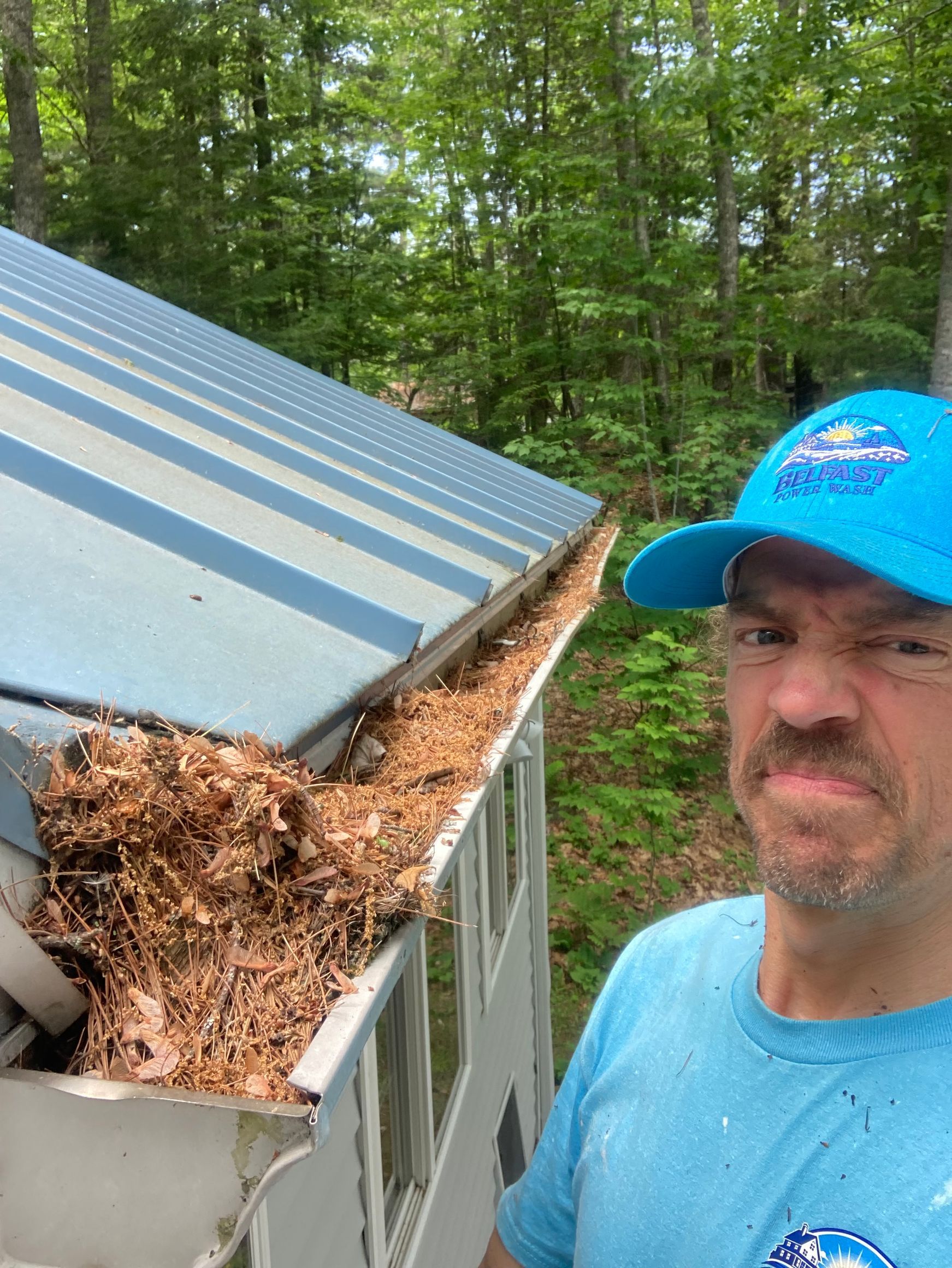 Man standing beside overflowing debris in a dumpster with a slanted metal lid, wooded background