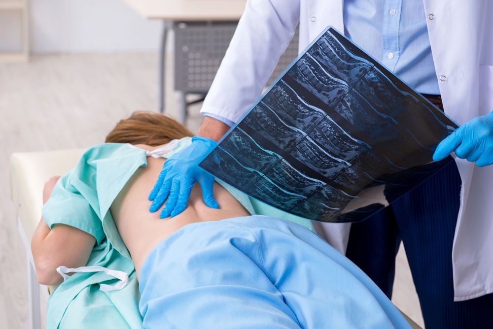 In un ambulatorio, un medico in camice bianco e guanti blu esamina la schiena di un paziente tenendo in mano una radiografia della colonna vertebrale.