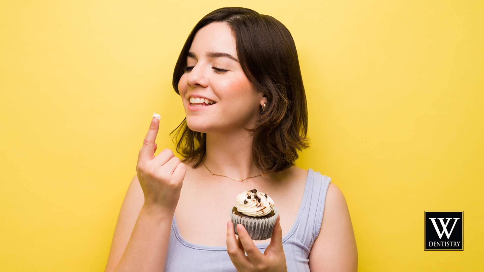 Woman with cupcake, tasting frosting from her finger, smiling, yellow background.