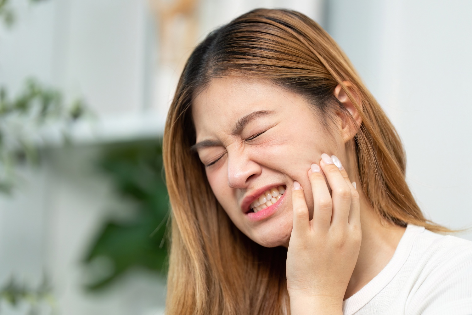 Woman grimacing and holding her cheek, suggesting tooth pain indoors