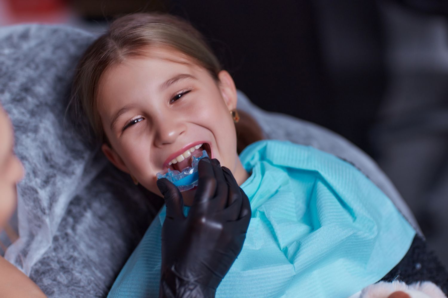 Child smiling while wearing a blue dental bib during a teeth cleaning with a black-gloved hand.