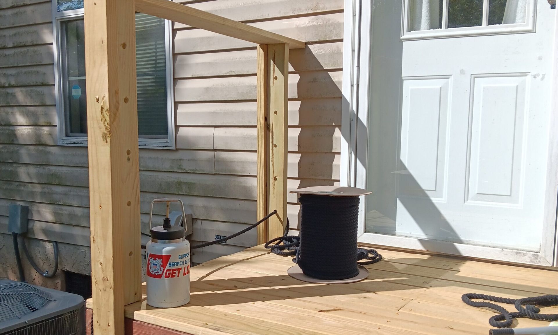 Wooden porch with railing under construction, paint bucket and black spool near a white door.