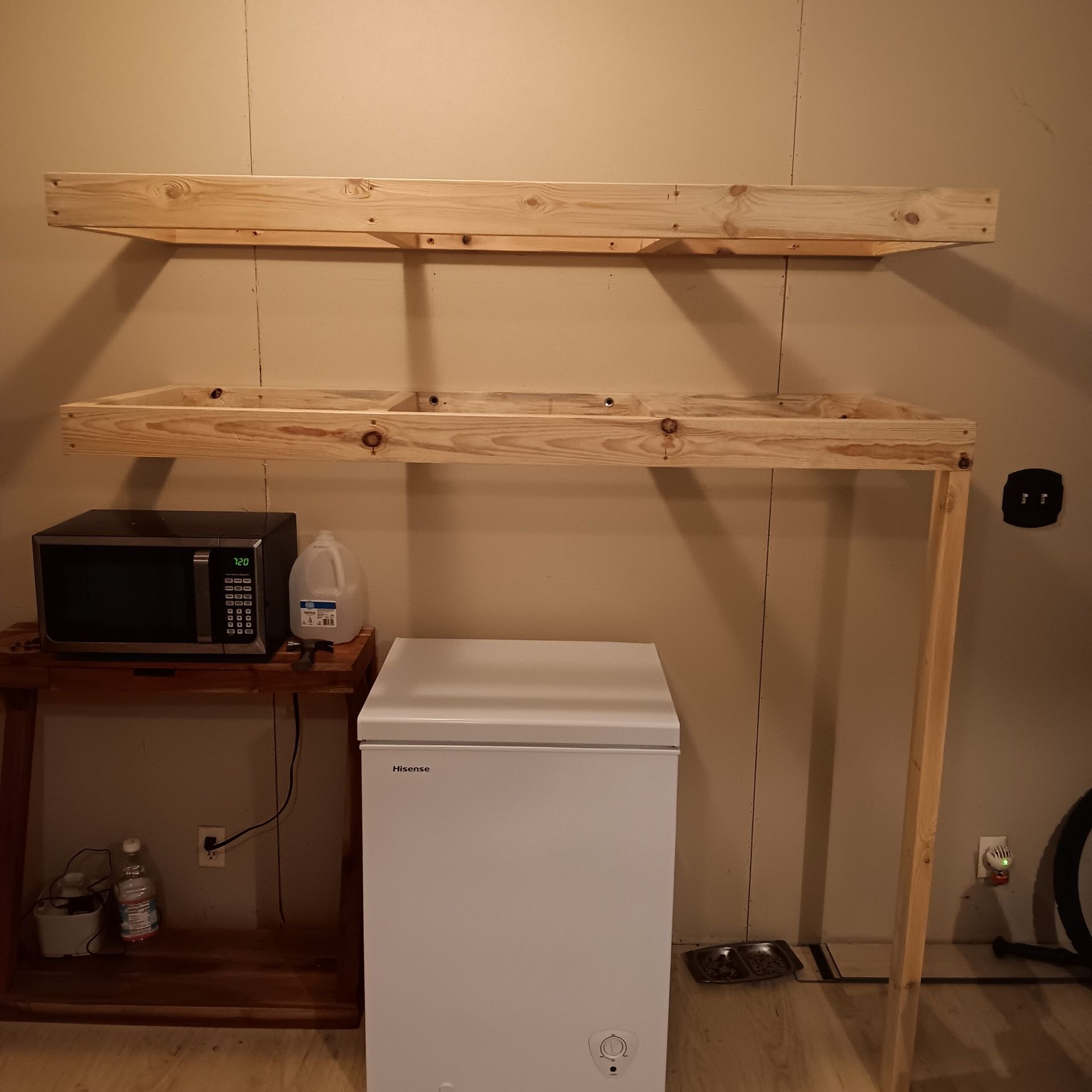 Unfinished wooden shelves above a white mini fridge in a small utility room with a microwave on a side table