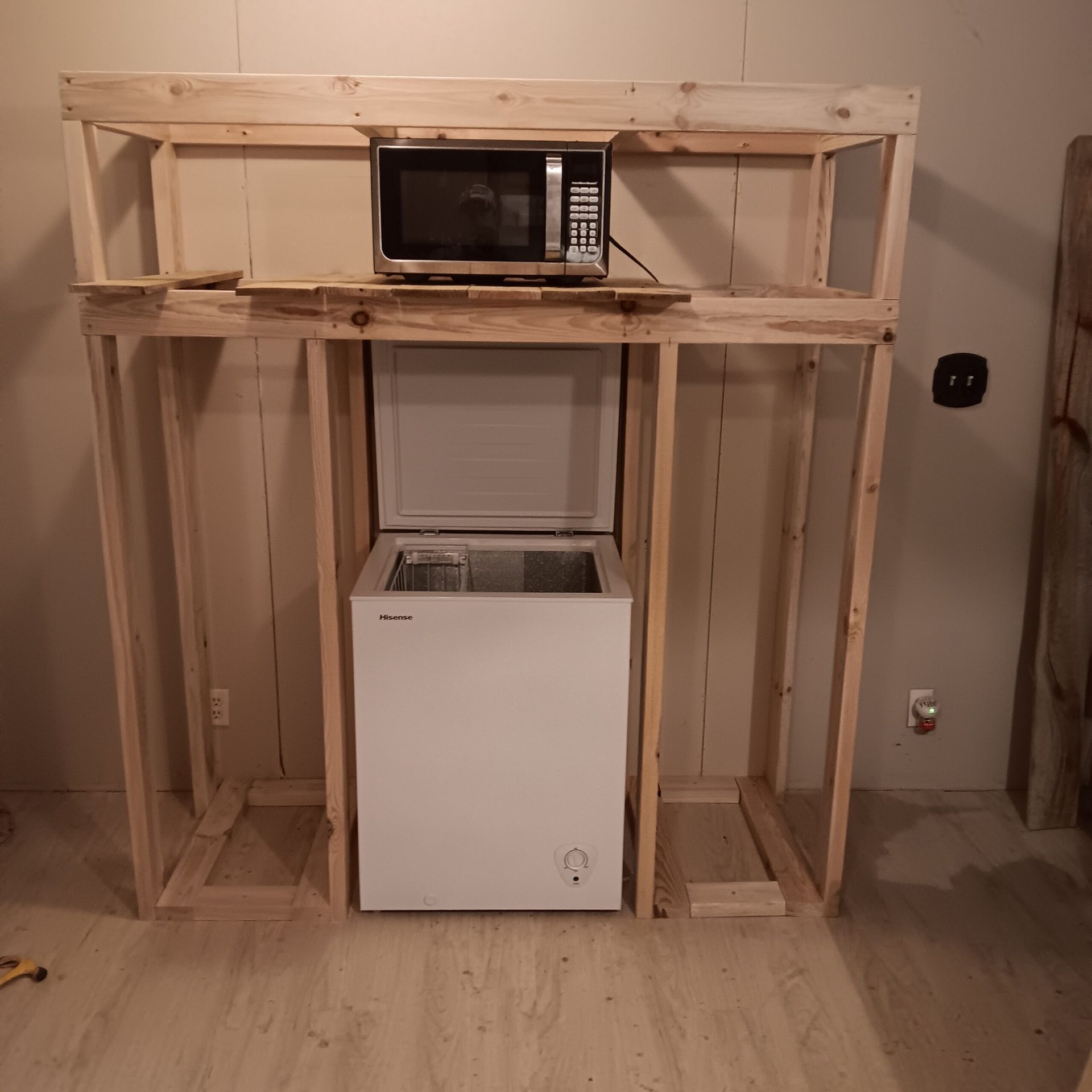 Microwave on a wooden shelf above a small white fridge in a bare room