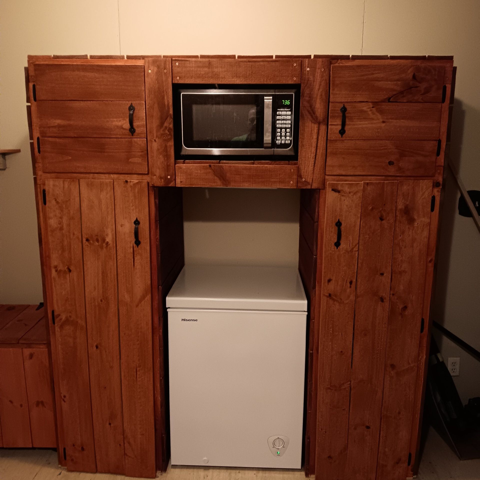 Wooden kitchen cabinet with built-in microwave above a white mini fridge on a beige wall.