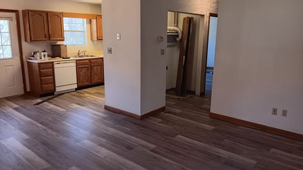 Empty kitchen and living area with wood floors and cabinets, seen from a corner doorway.