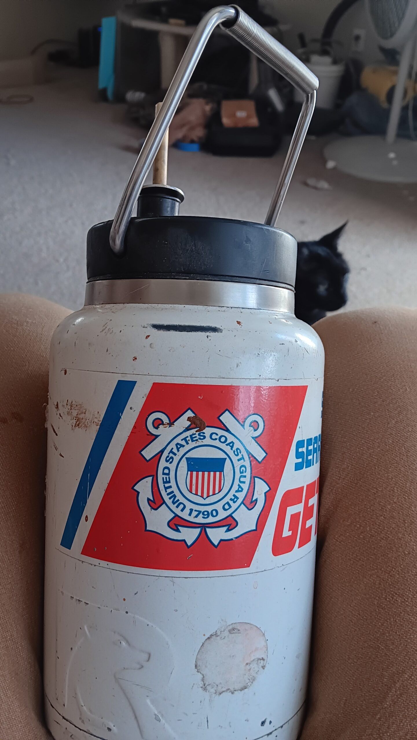 White metal canister with a handle and red logo label, sitting indoors on a floor.