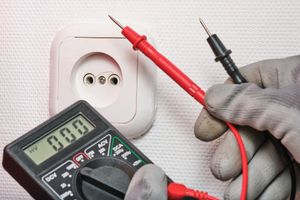 Hands testing an electrical wall outlet with a multimeter and red/black probes