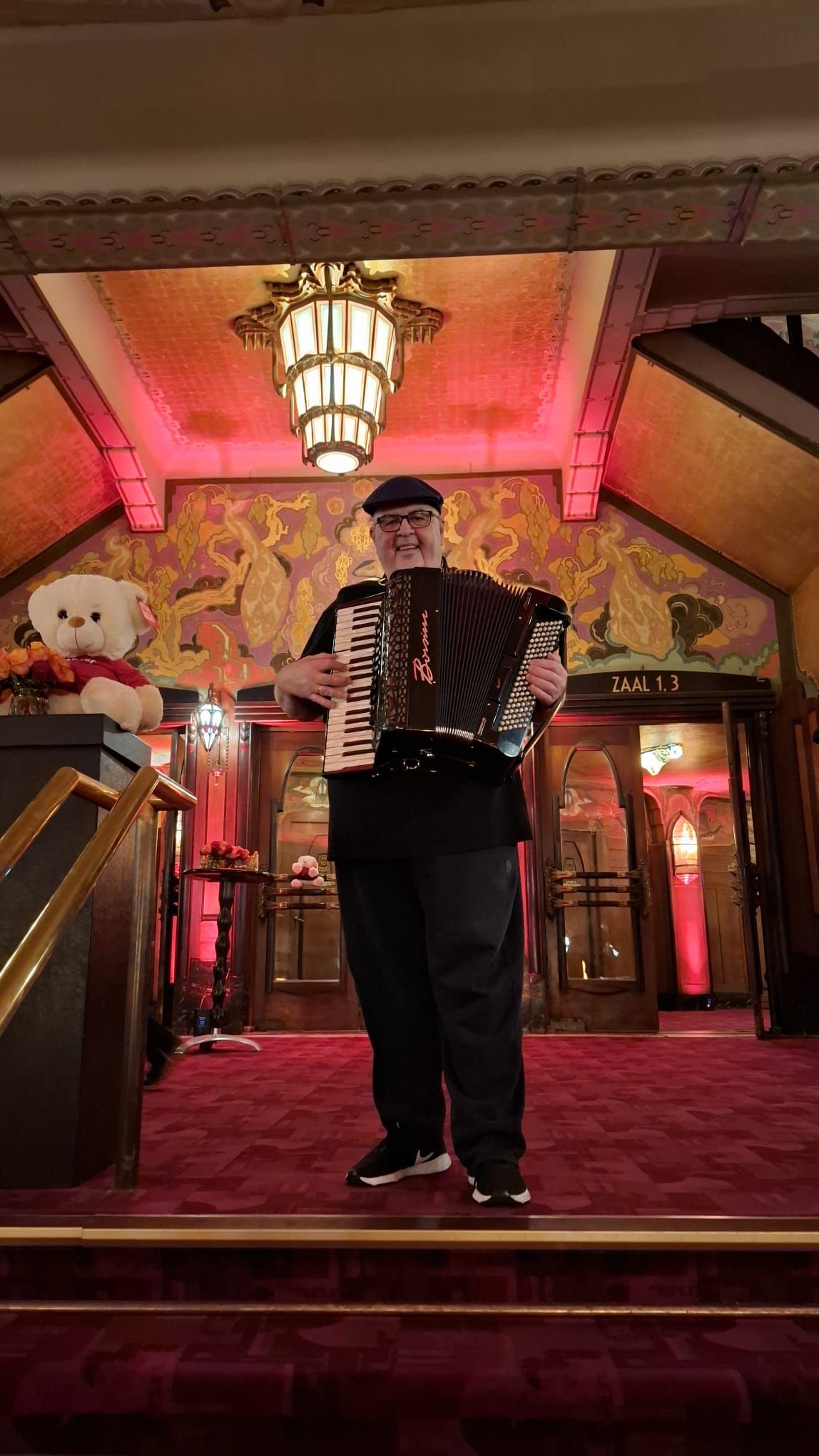 Een man speelt accordeon op een met rode loper bedekte trap in een warm verlicht, rijk versierd interieur.