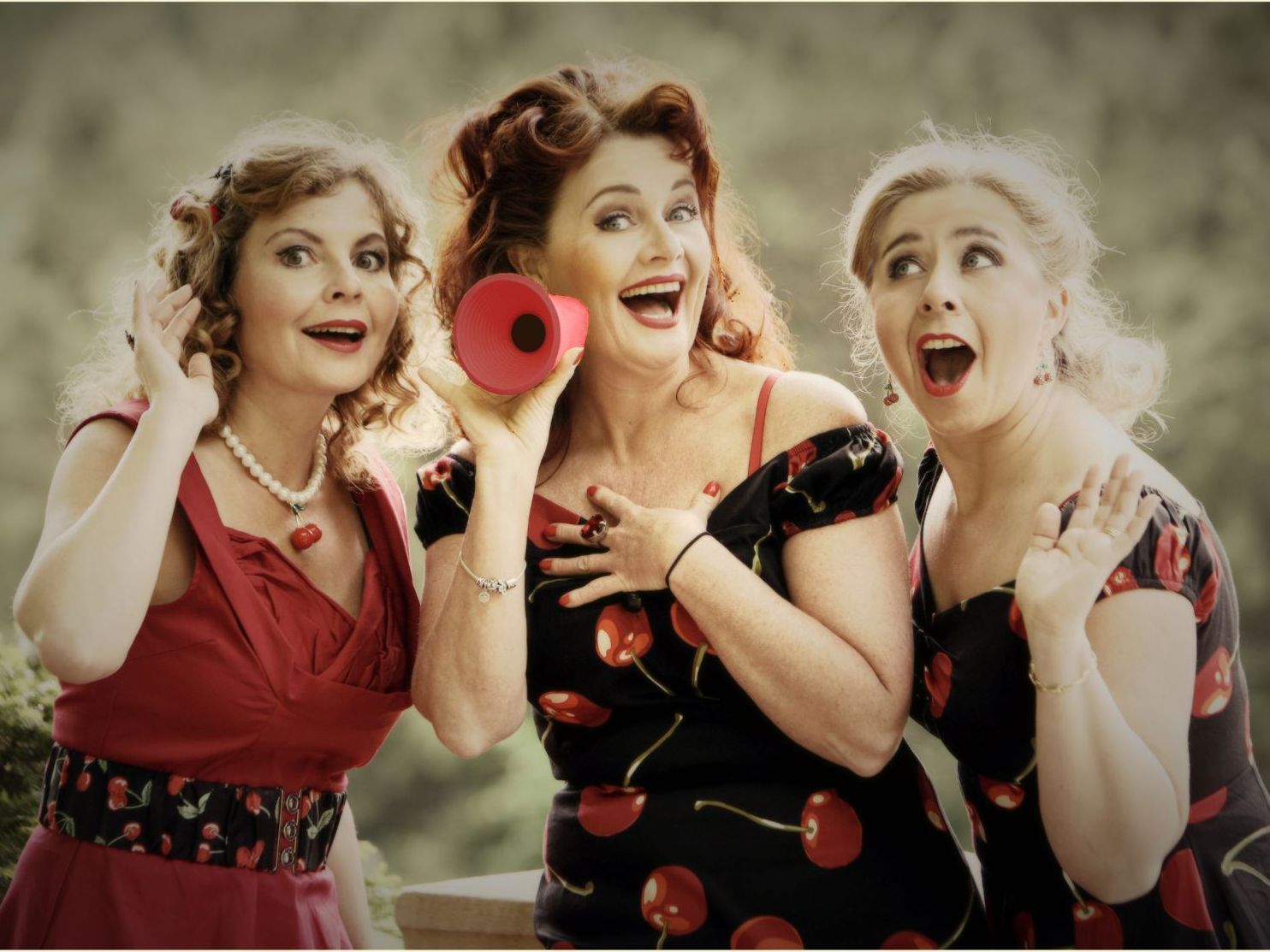 Drie vrouwen in rood-zwarte jurken glimlachen en zingen in een rode megafoon te midden van zwevende bubbels.