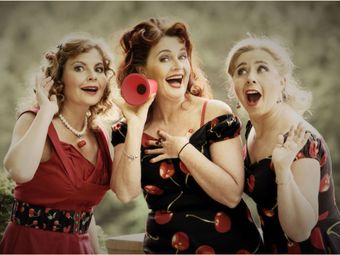Drie vrouwen in rood-zwarte jurken glimlachen en zingen in een rode megafoon te midden van zwevende bubbels.