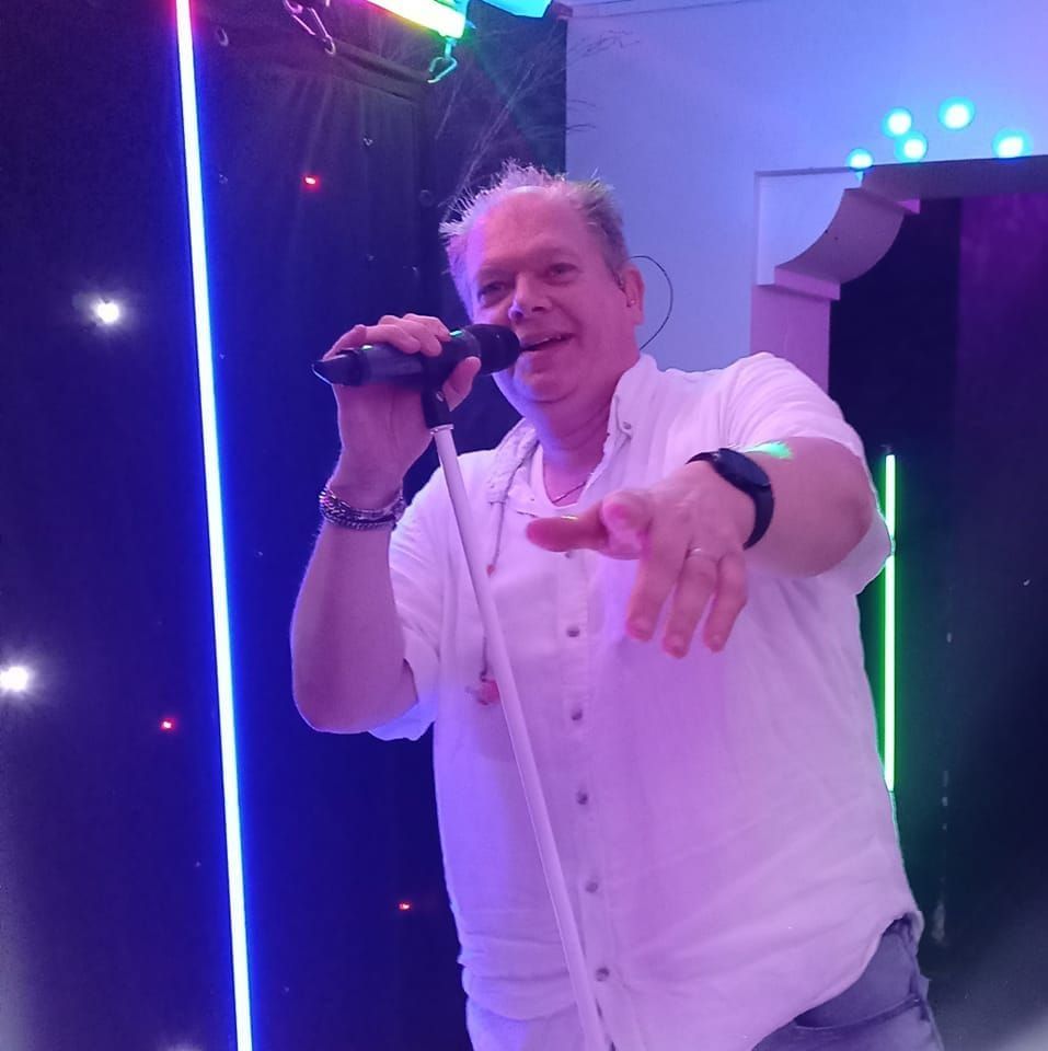 Zangeres in een wit shirt met een microfoon op een podium verlicht door neonlicht.