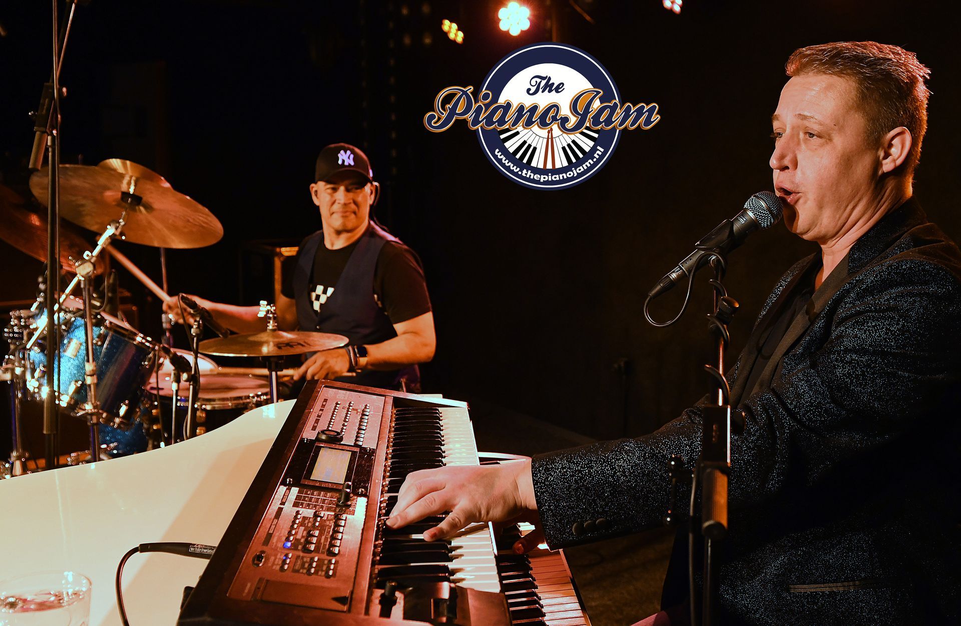 Muzikanten treden op het podium op, met de toetsenist op de voorgrond en de drummer erachter, onder de warme podiumverlichting.