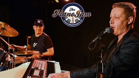 Zanger aan de microfoon en drummer die optreden op het podium onder warme verlichting met een bandlogo op de achtergrond.