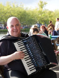 Een man speelt buiten op een picknicktafel op een zwarte accordeon, met mensen en bomen wazig op de achtergrond.