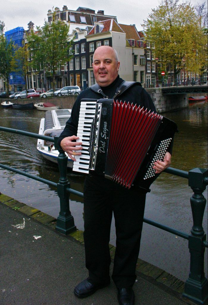 Een man speelt op een rode accordeon naast een kanaal in een regenachtige straat, met gebouwen en bomen op de achtergrond.