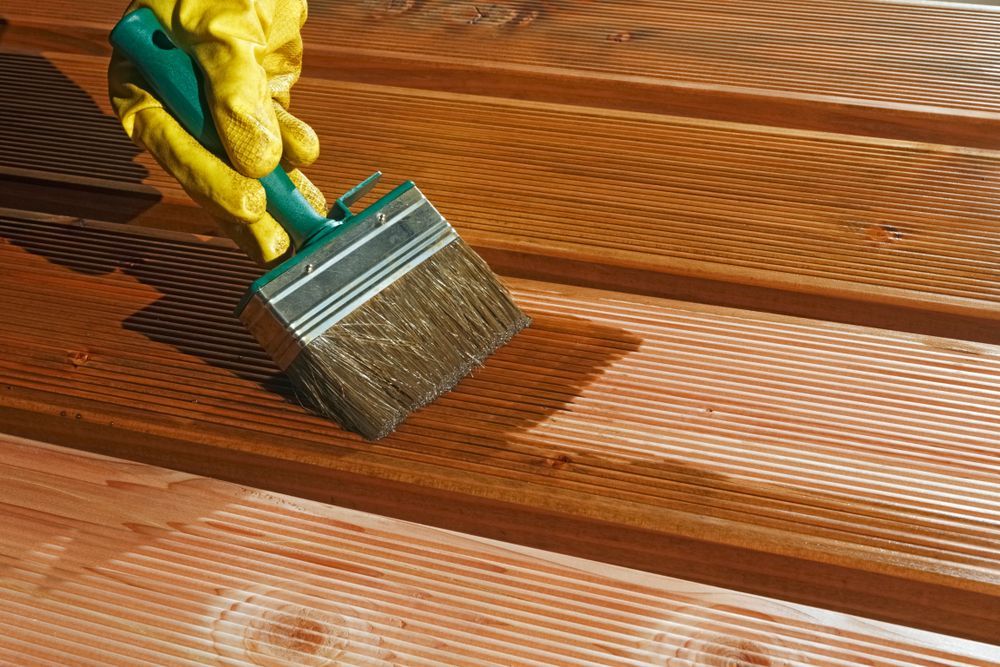 A hand in a yellow glove uses a brush to apply a brown wood stain or finish to ribbed wooden decking.