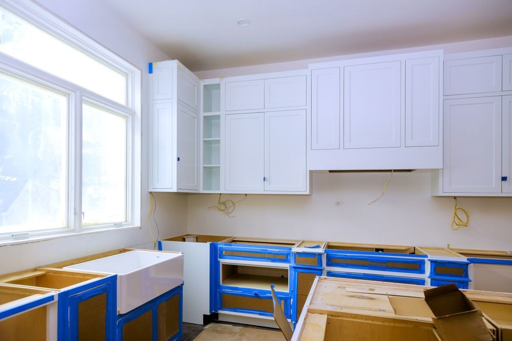 A kitchen undergoing renovation with white cabinets, a farmhouse sink, and blue painter's tape on unfinished countertops.