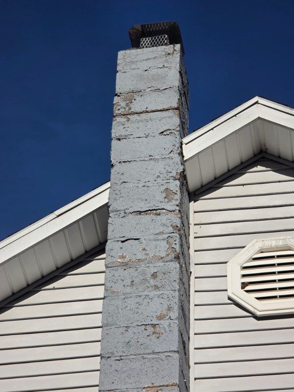 A gray concrete block chimney against a clear blue sky, situated next to the vinyl-sided wall of a house.
