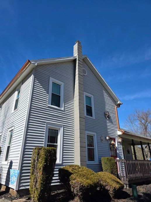A light gray two-story house with a prominent exterior chimney, a front porch, and trimmed bushes under a bright blue sky.