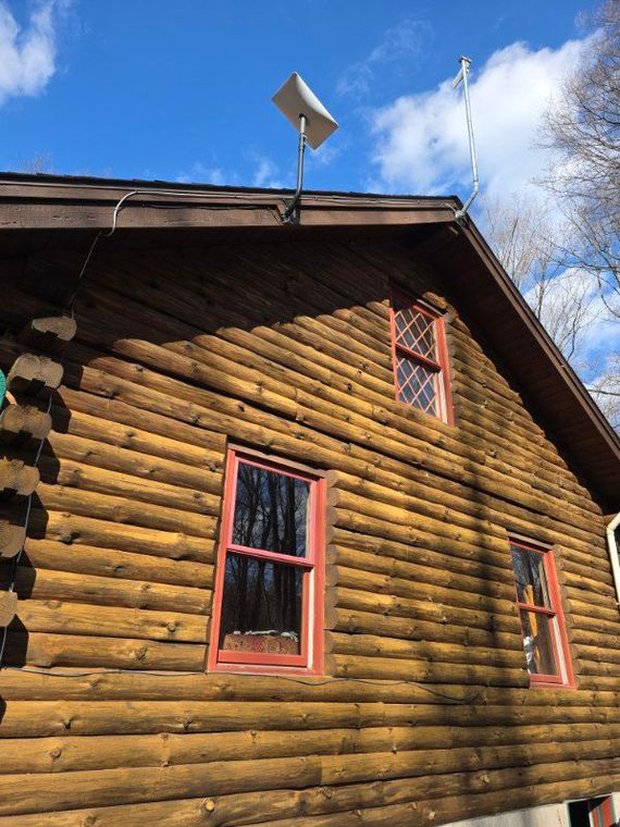A side view of a brown log cabin with red-trimmed windows and a satellite internet dish mounted on the roof.