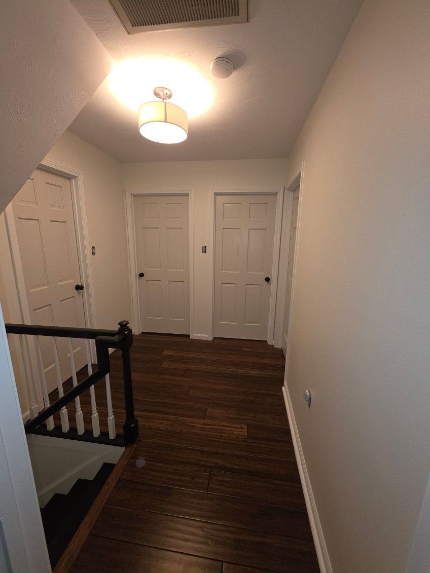 A hallway with dark wood floors, white walls, three white doors, and a staircase with a black railing.